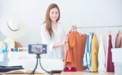 Beautiful asian woman blogger showing clothes in front of camera to recording vlog video live streaming at her shop.Business online influencer on social media concept.